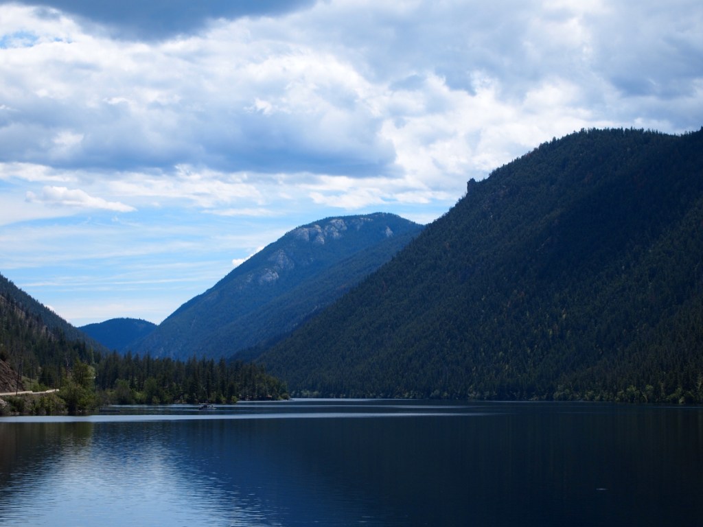 A placid mountain lake with steep woodland sloping directly into the water. At the left of the photo, a narrow road winds along the lakeshore.